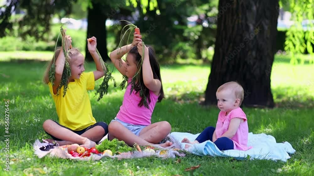 Three little girls on the lawn surrounded by various vegetables and fruits are playing with raw carrots Three little girls on the lawn surrounded by various vegetables and fruits are playing with raw carrots