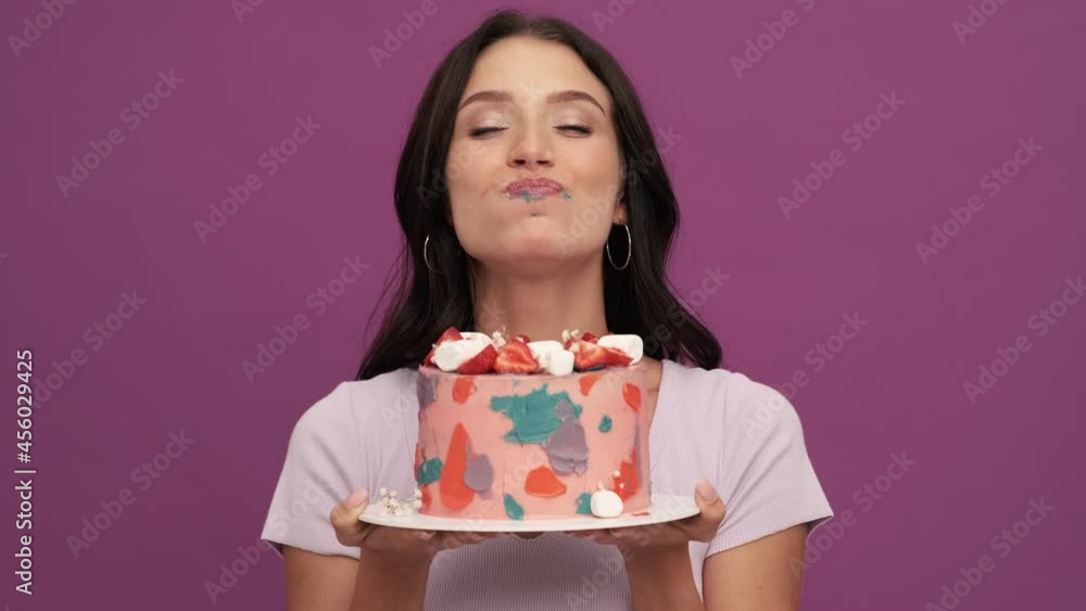 A smiling woman eating a birthday cake standing isolated over violet background in the studio A smiling woman eating a birthday cake standing isolated over violet background in the studio