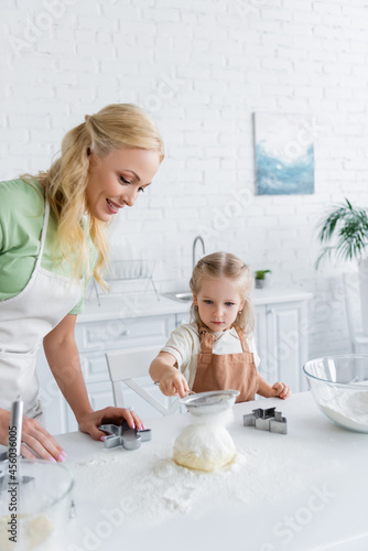 child sifting flour on raw dough while helping mom in kitchen