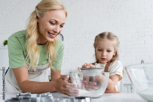 cheerful woman holding bowl with flour near little daughter with sieve