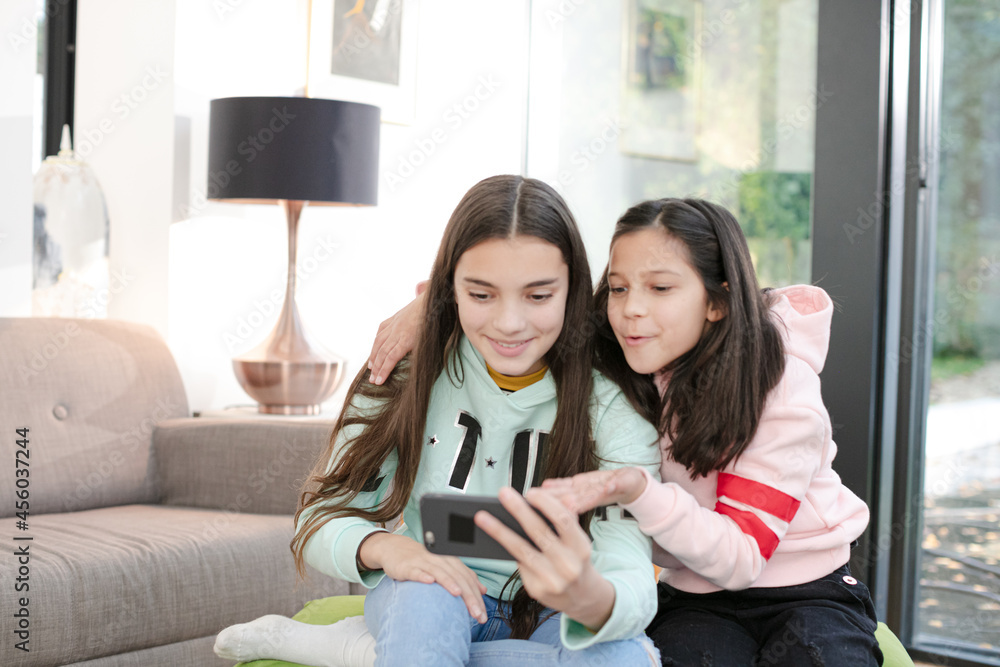 Playful sisters taking selfie with smart phone in living room Playful sisters taking selfie with smart phone in living room