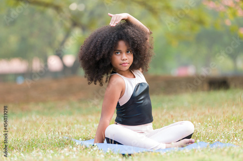 Happy African American little girl practicing yoga and stretching her hands on exercise mat outdoor in the park