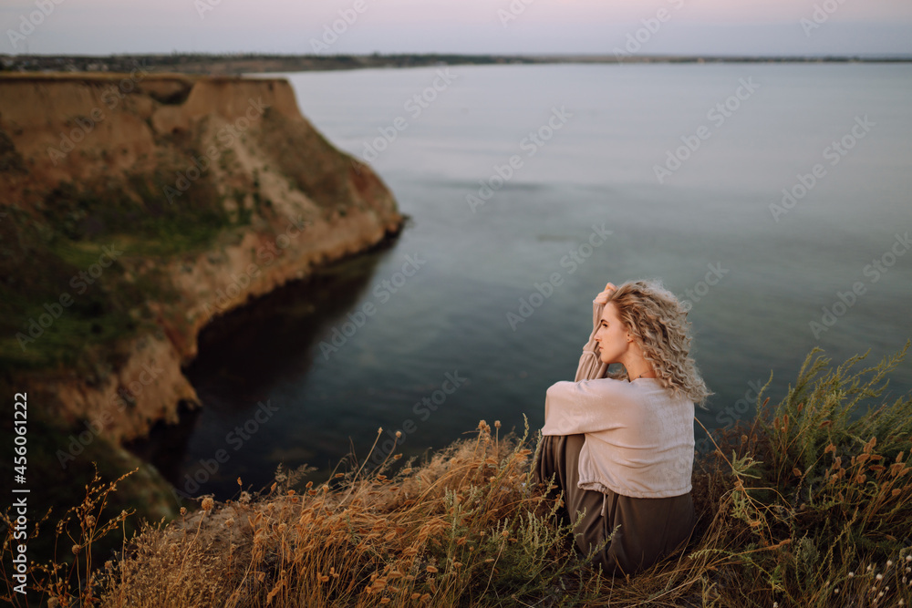 Fototapeta premium Stylish woman enjoys autumn nature at the edge of a rock above the sea, with blue sky and sea background. People, freedom, lifestyle, travel and vacations concept.