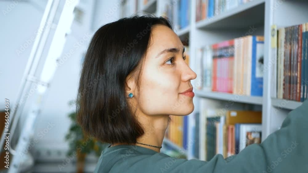 Young attractive female student selects an interesting book on the bookshelves in a public library.Close up portrait of a young attractive female student she is looking for the right book on the Young attractive female student selects an interesting book on the bookshelves in a public library.Close up portrait of a young attractive female student she is looking for the right book on the