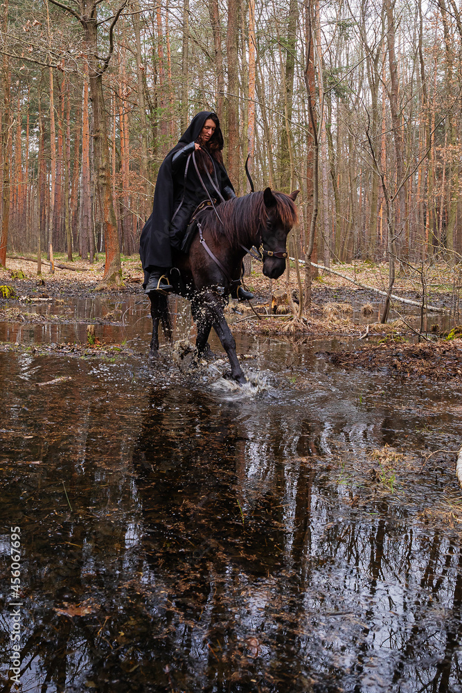 a woman in a historical costume with a bow and arrows riding a horse through a forest swamp