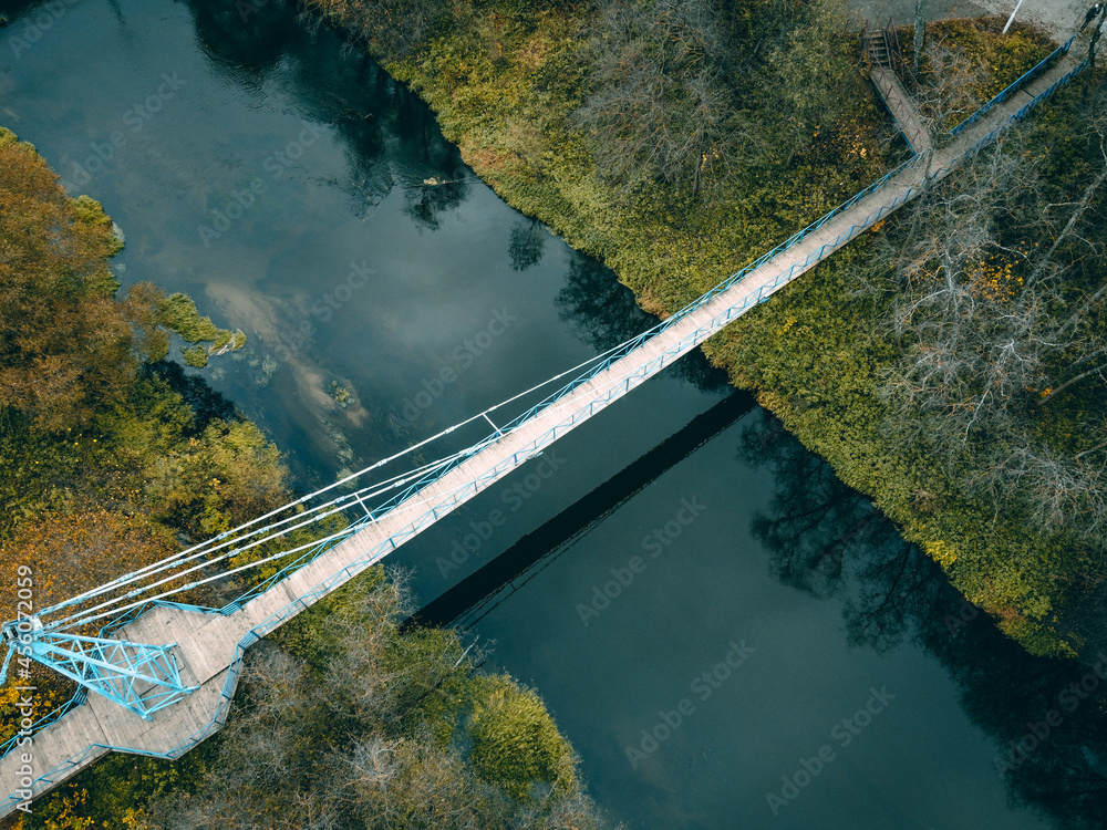 Fototapeta premium a small cable-stayed bluish-white bridge with a wooden floor over a narrow river, green grass and bushes on the sides
