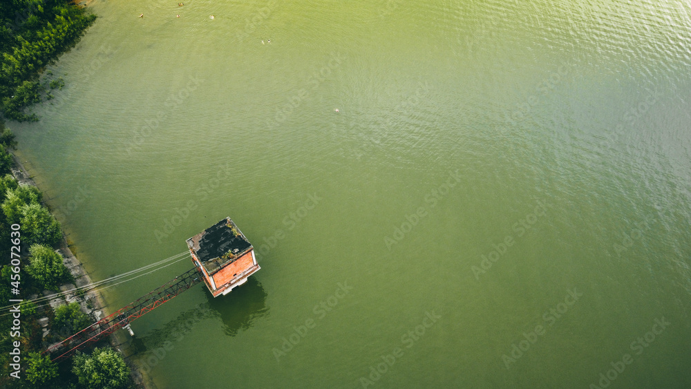 Obraz premium a marsh-colored lake, in the corner there are green bushes on concrete slabs, a small red-brick building with a black roof stands on the water across a small iron bridge