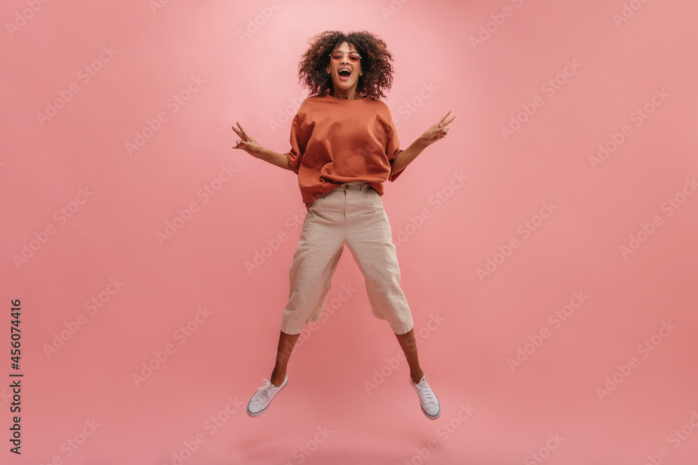 full-length image of happy African woman jumping against background of wall of pastel pink color. young brunette in sunglasses with curly hair shows peace sign with two hands. full-length image of happy African woman jumping against background of wall of pastel pink color. young brunette in sunglasses with curly hair shows peace sign with two hands.
