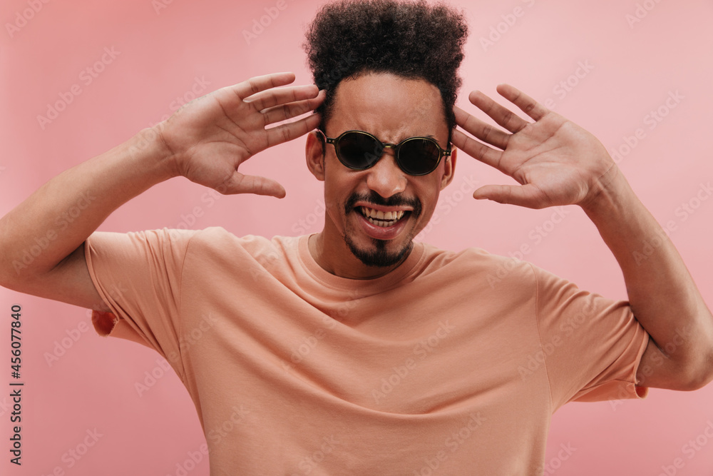 contented young bearded dark-skinned brunette showing his perfect white teeth. holding hands raised at head level on isolated studio background. man in fashionable sunglasses. contented young bearded dark-skinned brunette showing his perfect white teeth. holding hands raised at head level on isolated studio background. man in fashionable sunglasses.
