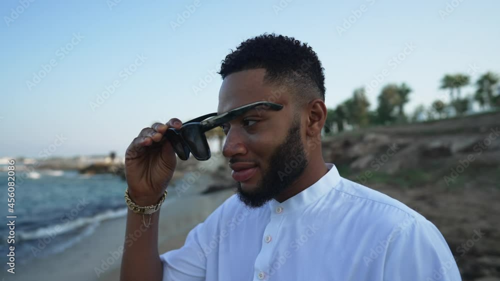 Young bearded handsome LGBT man putting on sunglasses in slow motion smiling. Portrait of happy African American gay person resting at Mediterranean summer resort outdoors. Diversity and leisure Young bearded handsome LGBT man putting on sunglasses in slow motion smiling. Portrait of happy African American gay person resting at Mediterranean summer resort outdoors. Diversity and leisure