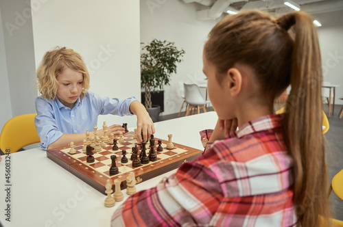 Serious little caucasian boy making his move while playing chess with friend, sitting together at the table in school