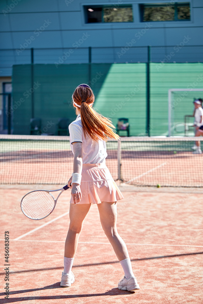 View From Back On Slim Female playing tennis and preparing for sports competition. Professional athlete in stylish uniform, in short skirt and cap. View from back