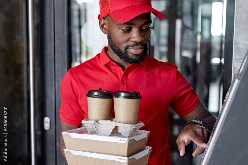 Courier holding takeaway food and drinks near house outdoors. Delivery service during quarantine due to Covid-19 outbreak. Black guy in red cap and uniform holding orders for clients or customers