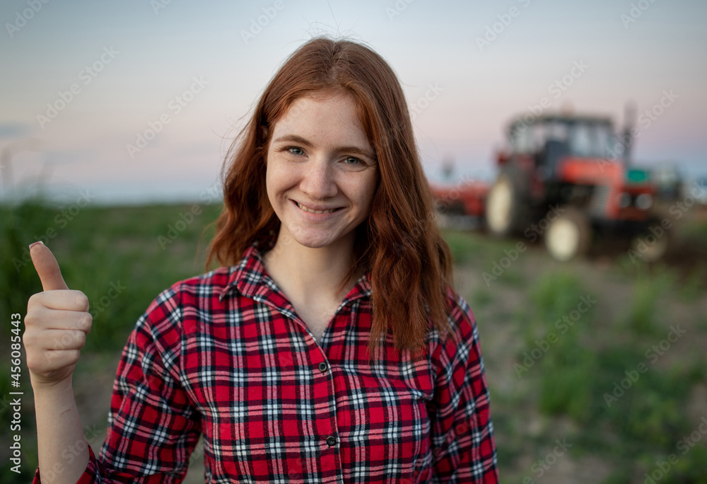 Portrait of young female farmer showing thumbs up in field with tractor in background