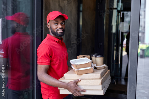 Positive Male courier with cardboard boxes delivering coffee cups and pizza to customer while standing outdoors of modern house, black man is looking happy, smiling, wearing red uniform, cap