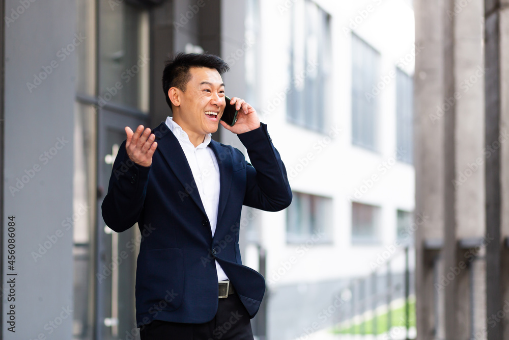 Asian businessman talking on the phone cheerful smiling reports good news near the office center outside Asian businessman talking on the phone cheerful smiling reports good news near the office center outside