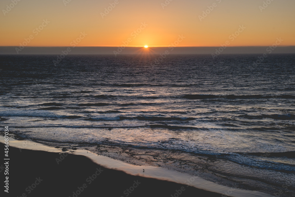 Panoramic view of the pacific ocean and beautiful sunset in the horizon with sun and rays in the center and the shore and beach with birds silhouettes