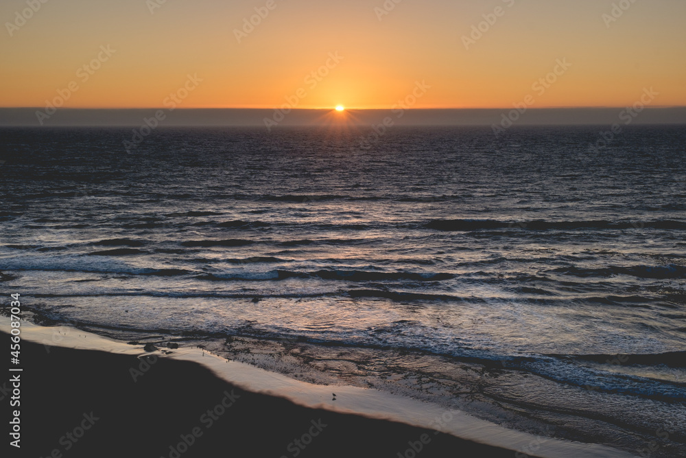 Fototapeta premium Panoramic view of the pacific ocean and beautiful sunset in the horizon with sun and rays in the center and the shore and beach with birds silhouettes