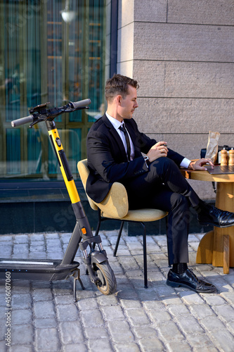 Young guy sit outdoors in cafe behind table using laptop, working online, side view. Guy in black suit sitting alone, using electro scooter, modern technologies concept. business people, lifestyle
