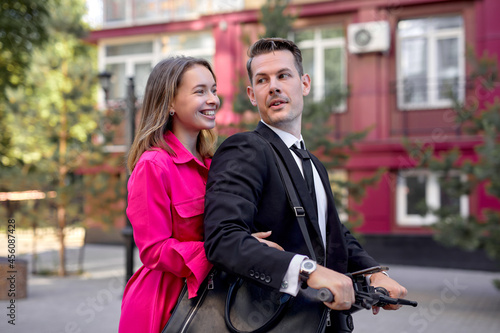married couple in stylish bright clothes driving electric scooter on city street, side view portrait of beautiful man and woman in suit and dress having fun, enjoying walk on scooter, have rest