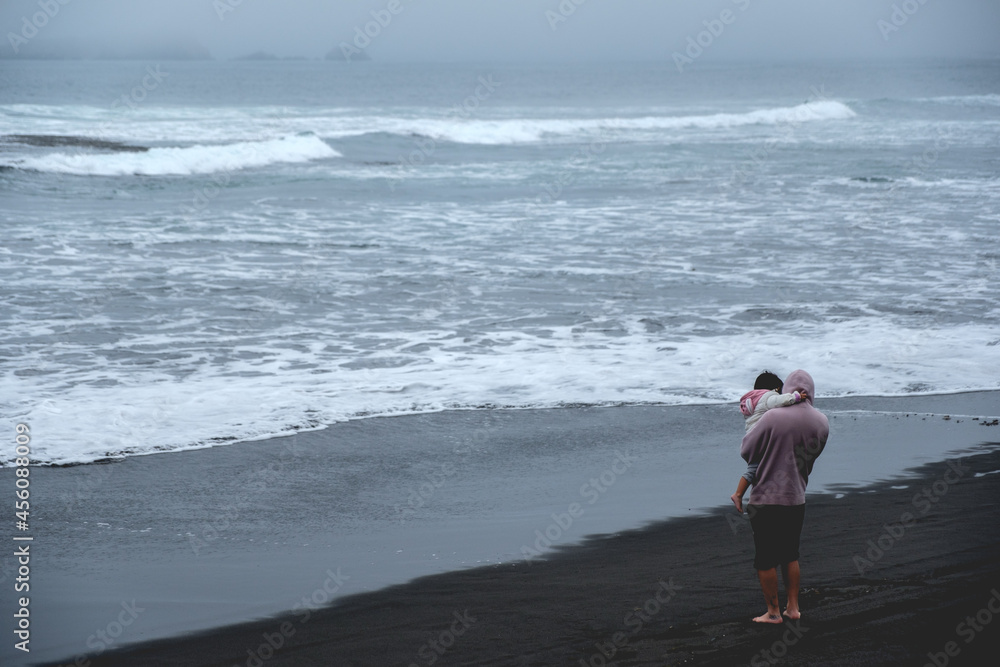 Dad and daughter (family) in front of a panoramic view of the pacific ocean with waves, the horizon with cloudy sky and the shore and beach Dad and daughter (family) in front of a panoramic view of the pacific ocean with waves, the horizon with cloudy sky and the shore and beach