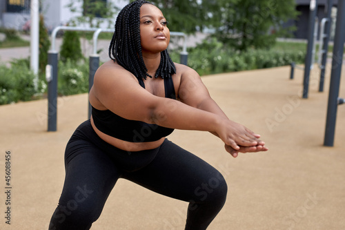 Fat african woman is engaged in fitness on sports ground outdoors, squatting. Beautiful young afro black overweight woman crouches outdoors. Exercises on muscles of buttocks, side view