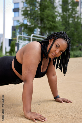 Beautiful young fat african woman wearing black sportswear working out outdoors, doing yoga or pilates exercise. Push ups or press ups, Plank, phalankasana pose. Side view, look at camera