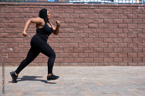 Side View On Confident Nice Obese Woman In Sportive Clothes Jogging, Running Forward, Training In City Street, Concentrated On Sport Jogging, Lead Healthy Lifestyle. Weight Loss Concept. Copy Space