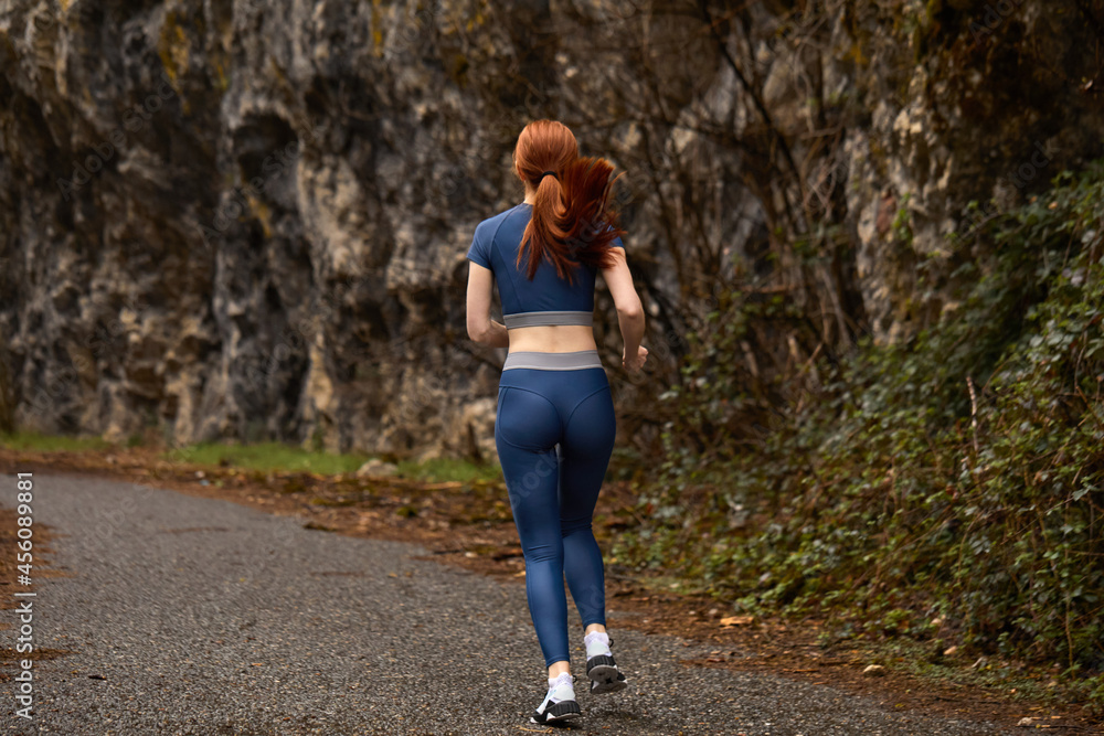 Rear view on redhead female jogging alone in nature, in blue sportive clothes, running, preparing for competition, outdoors. active and healthy athlete lady engaged in sport, lead healthy lifestyle