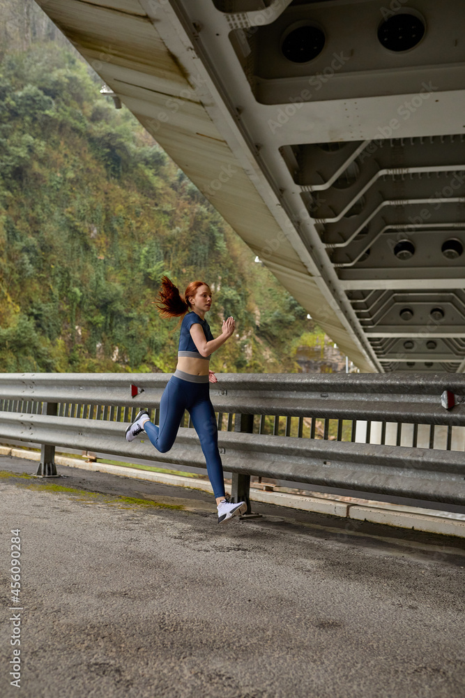 Beautiful Caucasian Female In Sportive Clothes Jogging Alone On Fresh Air, On Bridge, Side View On Active Healthy Lady Motivated By Sport, Fitness. Redhead Woman Lead Healthy Lifestyle