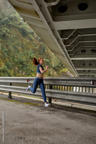 Beautiful Caucasian Female In Sportive Clothes Jogging Alone On Fresh Air, On Bridge, Side View On Active Healthy Lady Motivated By Sport, Fitness. Redhead Woman Lead Healthy Lifestyle