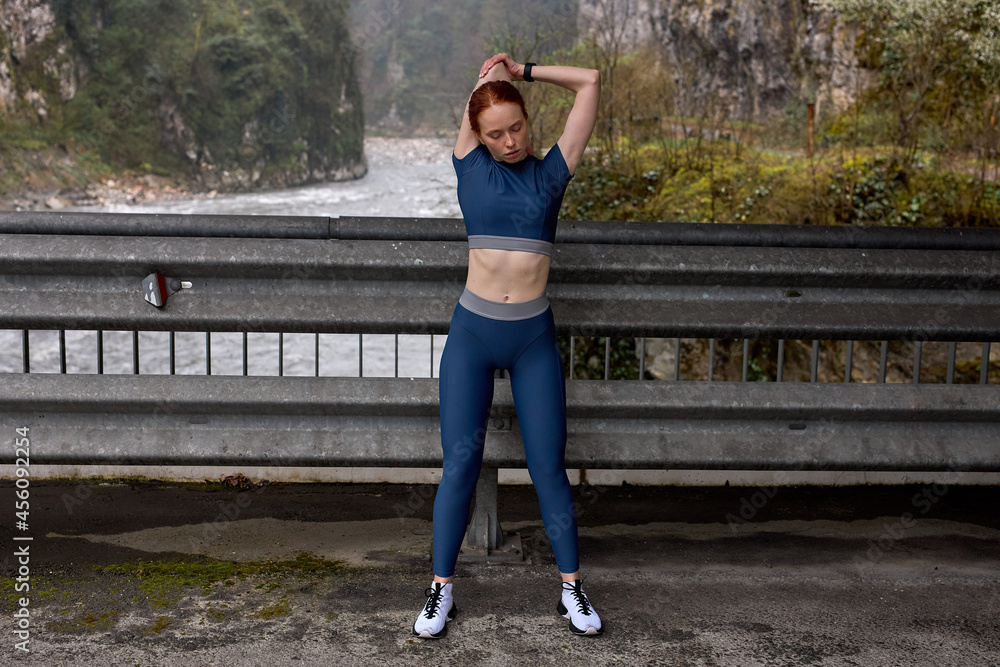 Slim fitness woman runner relaxing after outdoor running and working out. An attractive redhead female runner stretching before workout, in blue sportive outfit. Copy space, portrait. Sport concept