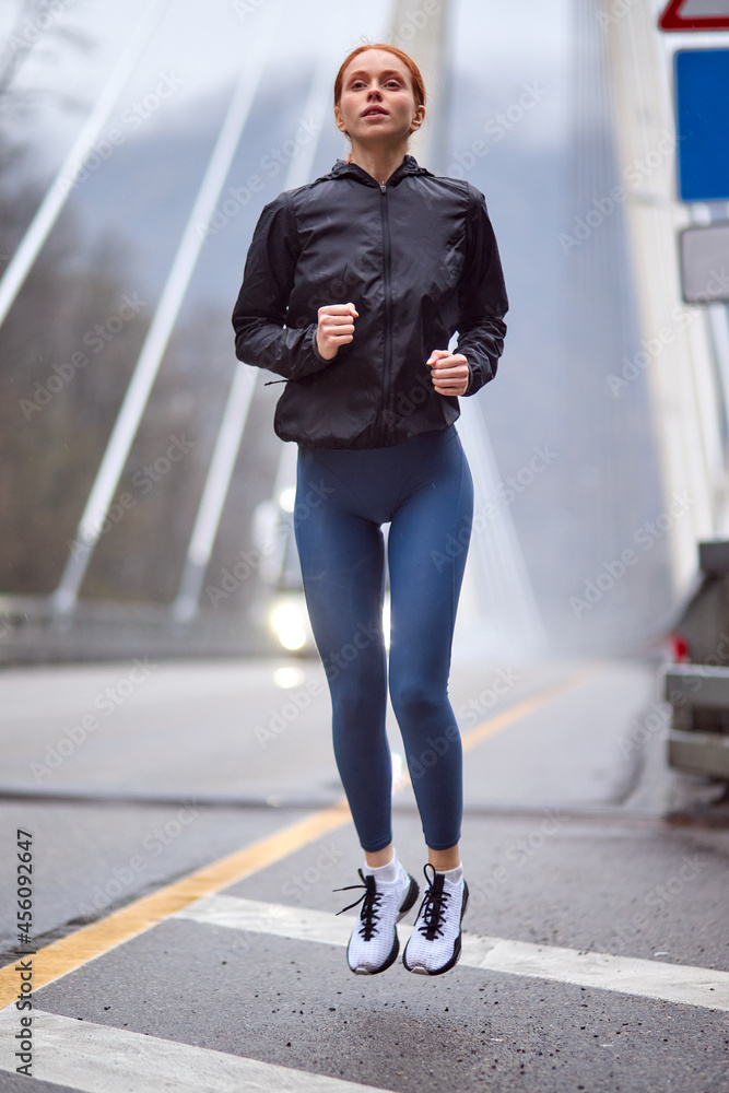 Foto de Portrait of confident female runner jogger training outdoors on  bridge alone, doing exercises, in sportive outfit, in the morning, cold  day. workout, sport, jogging, healthy lifestyle concept do Stock