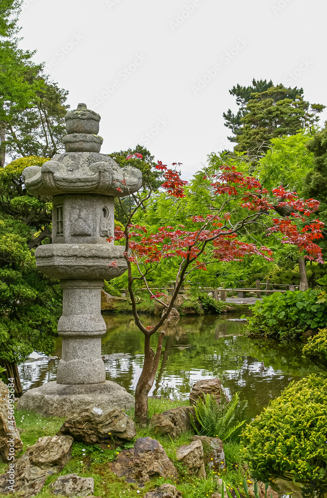 USA, California, San Francisco - May 19, 2008: Japanese Garden. Portrait of gray cut-stone lantern statue set near pond under silver sky with different shades of green and red foliage.