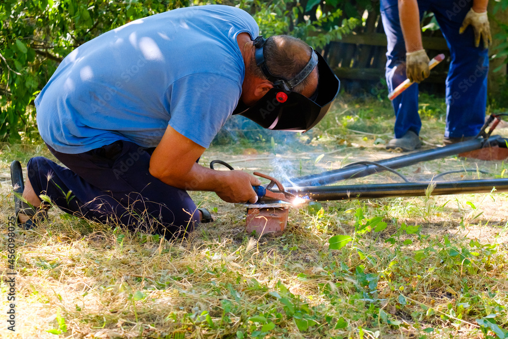 A worker wearing a protective mask welds metal pipes with electric arc welding on a sunny summer day. A worker wearing a protective mask welds metal pipes with electric arc welding on a sunny summer day.
