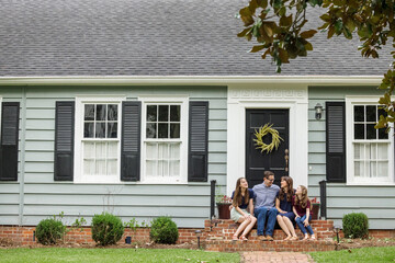 A family with a mother, father, and two daughters sitting outside on the brick steps of a front porch of a small blue cottage house