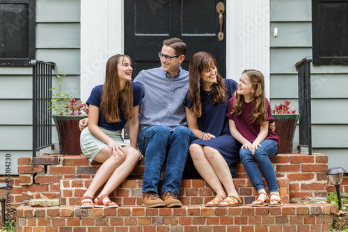 A family with a mother, father, and two daughters sitting outside on the brick porch of a small blue cottage house