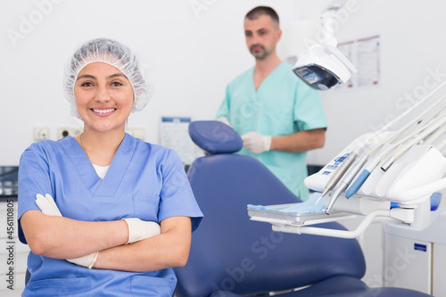 Cheerful latin female dentist in uniform with crossed hands sitting near medical chair