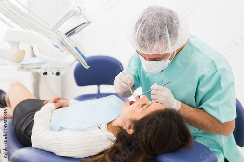 The dentist in face mask inspects female patient teeth with mirror and probe