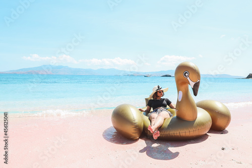 Asian woman in summer hat and sunglasses relaxing on inflatable swan in pink sandy beach