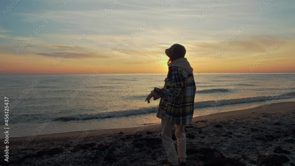 Rear view of woman wearing warm outwear dancing at sunset near the seaside. Silhouette of a woman dancing on the beach during an amazing sunset. Rear view of woman wearing warm outwear dancing at sunset near the seaside. Silhouette of a woman dancing on the beach during an amazing sunset.