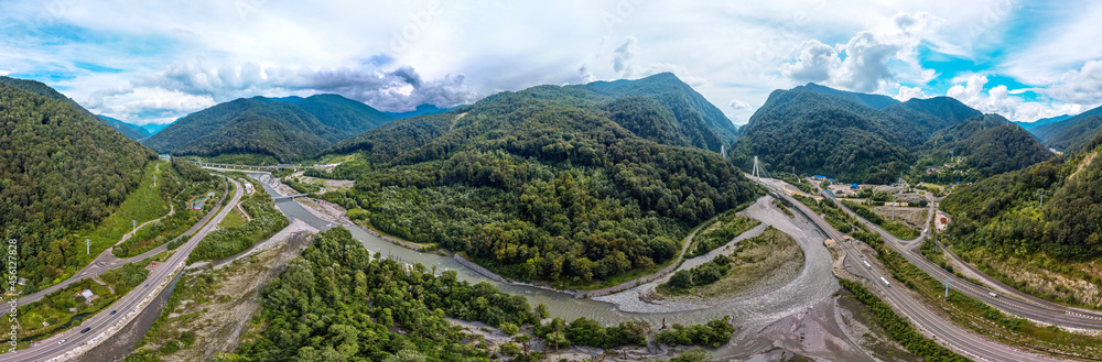 a large aerial panorama of the valley of the mountain river Mzymta, surrounded by highways and forested green mountains of the Caucasus. Cable-stayed bridge over the river entering the tunnel