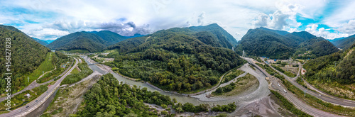 a large aerial panorama of the valley of the mountain river Mzymta, surrounded by highways and forested green mountains of the Caucasus. Cable-stayed bridge over the river entering the tunnel