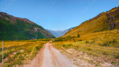 Path through mountains. Trekking mountain trail. Bright panoramic alpine landscape with dirt road among grasses in highlands. Pathway uphill. Way up mountainside.
