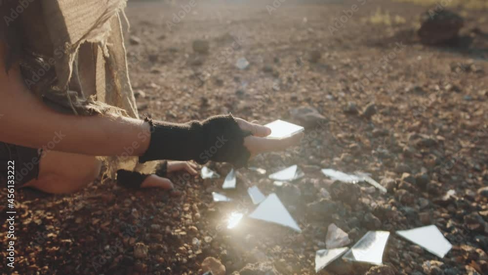 Handheld camera shot of young woman in shabby clothes looking at pieces of broken mirror in dystopian world