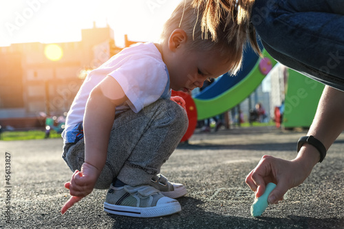 The son and mother are painting with chalk on the playyard. The girl is writing whiting on the asphalt, and the squatting boy is watching photo