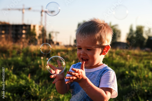 Smiling boy with blond hair catches soap bubbles against the background of a green field. A caucasian child is playing in the city with a view of the construction site and the cranes.