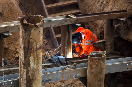 A man in uniform and mask is welding a water pipe. Construction of metal pipes and a builder