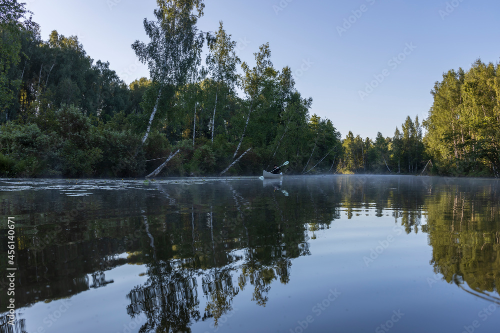 Fototapeta premium A man in a kayak floats down the river. Picturesque landscape in the morning on the river. The sun's rays touch the treetops. Kayaking early in the morning on a calm river in the summer.