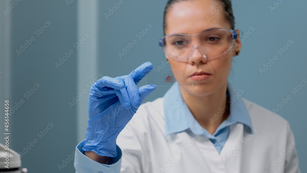 Fototapeta premium Close up of biology doctor looking at blood test on glass tray in science laboratory. Scientist woman wearing glasses and gloves for protection at clinic analyzing dna sample for experiment